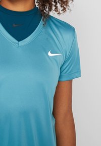 Turkos Nike sportshirt med V-hals, korta ärmar och en vit logotyp på vänster bröst. Lager ovanpå ett mörkblått baslager. Slät textur.