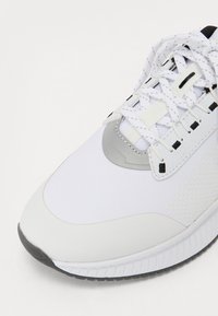 White athletic shoe with a textured surface, a silver accent, and black detailing; features laces and a cushioned sole for support.