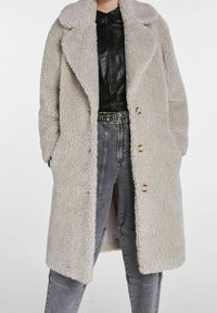 Cremefarvet faux shearling frakke med reverskrave, lange ærmer og tre knaplukninger. Lommer på begge sider, parret med grå jeans.