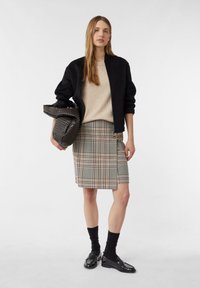 Zwarte bomberjack over een beige trui, gecombineerd met een geruite rok tot de knie. Houdt een donkere, geweven tas vast. Zwarte sokken en schoenen maken de look compleet.