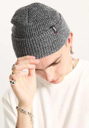 Bonnet tricoté gris avec une texture côtelée et une petite étiquette logo. Le tissu est extensible et ajusté, offrant de la chaleur.