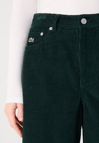 Dunkelgrüne Cordhose mit vertikaler Rippung, Knopfverschluss und kleinem gesticktem Logo auf der vorderen Tasche.