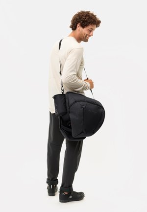 Sac à dos noir avec un tissu texturé et un motif géométrique ; il dispose de bretelles réglables et de poches extérieures. Le modèle porte une chemise claire et un pantalon foncé.