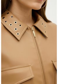 Tan kunstlæderjakke med nitter i guldfarve på kraven, lynlåslukning og to frontlommer med klap. Glat tekstur og struktureret design.