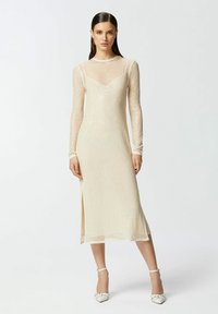 Haljina dugih rukava kremaste boje s uskim korzetom, prozirnim prekrivačem, suptilnim sjajem i bočnim prorezom. Izrađena od nježne, teksturirane tkanine.