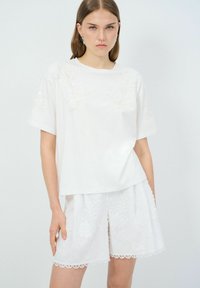 Top blanco de manga corta con diseños florales en relieve y corte recto, combinado con pantalones cortos blancos con adornos de encaje y bordados florales.