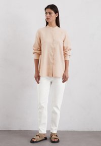 Lichtroze linnen shirt met een mandarinakraag, knopen aan de voorkant en lange mouwen, gecombineerd met witte jeans en bruine sandalen.