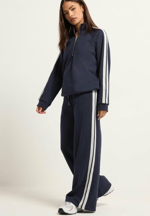 Femme debout portant un survêtement bleu marine avec des bandes blanches sur les manches et les jambes du pantalon, assorti à des baskets blanches, fond neutre.
