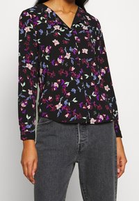 Blouse noire à fleurs avec manches longues, présentant des fleurs roses, blanches et bleues. Possède un col et un design à patte de boutonnage. Associée à un jean gris.