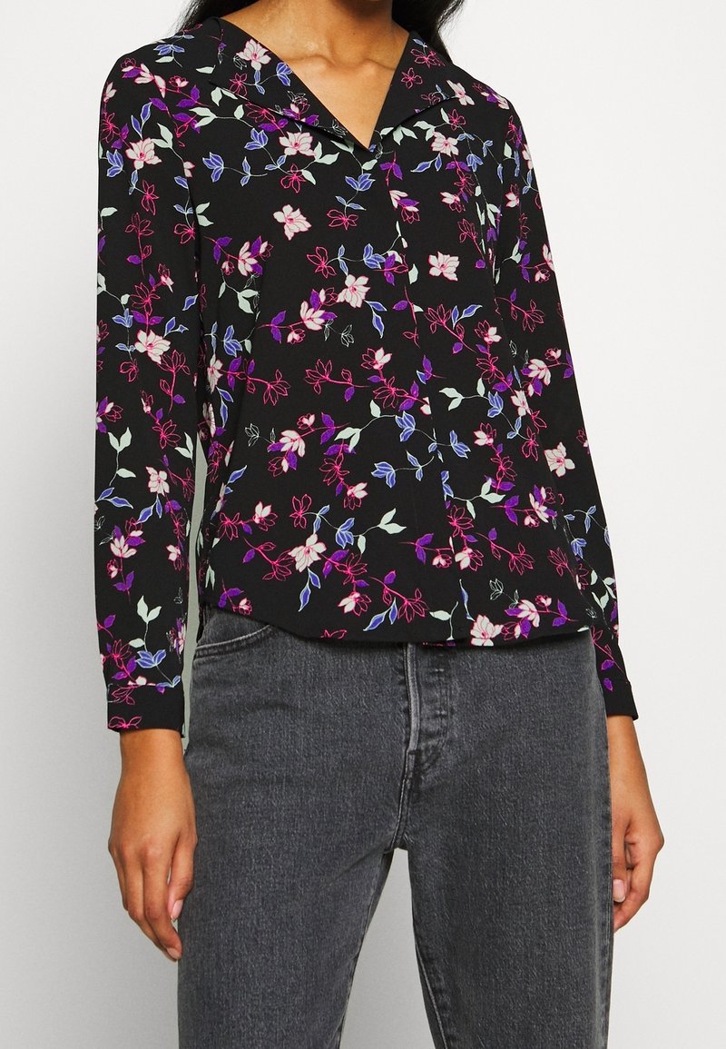 Blouse noire à fleurs avec manches longues, présentant des fleurs roses, blanches et bleues. Possède un col et un design à patte de boutonnage. Associée à un jean gris.