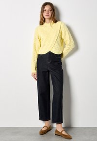 Żółty sweter z dzianiny warkoczowej z wysokim kołnierzem, zestawiony z ciemnymi dżinsami z wysokim stanem. Modelka ma na sobie brązowe zamszowe baleriny.