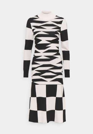 Robe en maille à manches longues présentant des motifs géométriques noirs et blancs, comprenant des zigzags et des motifs à carreaux, avec un col montant simulé.
