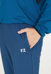 Blå idrottsbyxor med dragsko i midjan, sidoficka och vitt "FZ"-logotyp. Slät textur och lättmaterial.