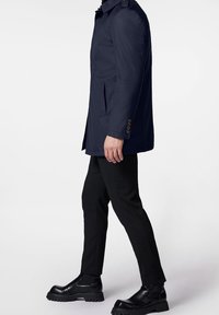 Manteau bleu marine au design structuré, avec des épaulettes, des poches latérales et des poignets boutonnés. Porté sur un pantalon noir et des bottes.