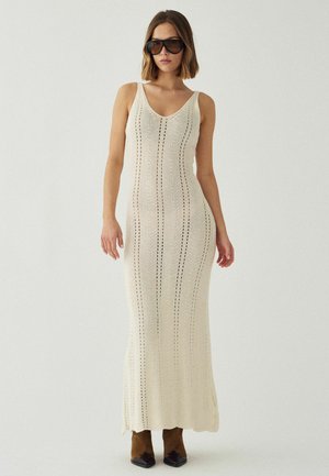 Nő, aki ujjatlan, hosszú krémszínű kötött ruhát visel, függőleges mintákkal, túlméretezett fekete napszemüvegben és barna, hegyes orrú csizmában áll.