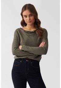 Pulover verde măsliniu cu decolteu rotund și mâneci lungi, textură moale, asortat cu blugi închiși la culoare, cu talie înaltă, prezentând cusături contrastante.