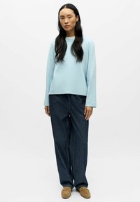 Jasnoniebieska, długorękawowa bluzka z miękkiego materiału, w zestawieniu z ciemnoniebieskimi, szerokimi dżinsami i beżowymi butami typu slip-on, minimalne detale.
