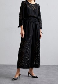 Musta kaksiosainen asu, jossa on löysä, läpikuultava paita, jossa on geometrija leikkuja, ja leveälahkeiset housut, joissa on joustava vyötärö. Yhdistetty mustiin korkokenkiin.