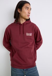Hanorac maron cu buzunar frontal, glugă cu șnur și logo brodat "Dickies Donut House" în alb și roz. Țesătură moale și texturată.