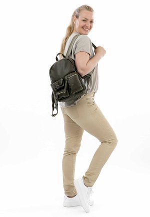 Sac à dos en cuir vert avec deux poches avant et des sangles réglables. Porté par une personne en pantalon beige et baskets blanches sur un fond blanc.