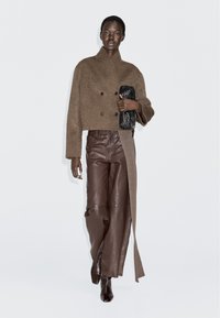 Model wearing brown cropped double-breasted wool jacket, wide-leg leather pants, brown boots, holding black textured clutch, walking on plain white background.