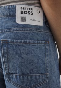 Jeanshosen-Hintertasche mit weißem Etikett mit der Aufschrift "Better Boss" und einem QR-Code, getragen von einer Person in einem gestreiften Hemd.