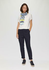 Witte t-shirt, marineblauwe broek op maat, zwarte sneakers met dikke beige zolen, en een kleurrijke sjaal met luipaardprint met gele en blauwe accenten.