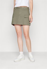 Jupe cargo vert olive avec deux poches avant, une coupe droite et une fermeture à bouton. Associée à des baskets blanches et des chaussettes de cheville.