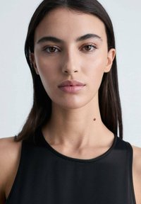 Black sleeveless top with a round neckline, smooth texture, worn by a model with straight dark hair and a beauty mark on the neck.