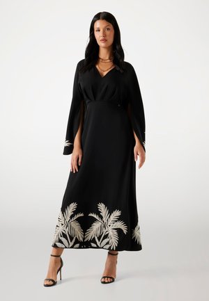 Femme portant une robe maxi noire à manches longues avec une broderie beige de feuilles de palmier à l'ourlet et aux manches, accompagnée de sandales noires à talons hauts.