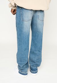 Jean en denim bleu clair avec une coupe ample, texture délavée, et deux poches arrière ; associé à des baskets blanches et un haut beige.