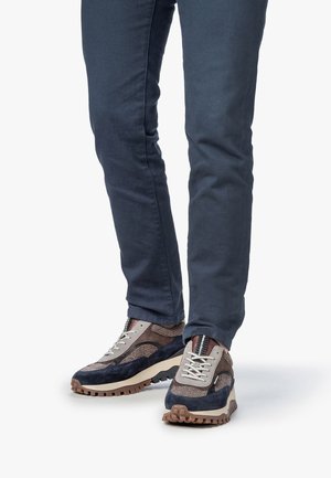 Beine in dunkelblauen, eng anliegenden Hosen und braunen, marineblauen und beigen lässigen Sneakers mit strukturierten Sohlen vor weißem Hintergrund.