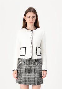 Veste blanche texturée avec des finitions noires, ornée de boutons ronds et de poches plaquées, associée à une jupe à carreaux noir et blanc.