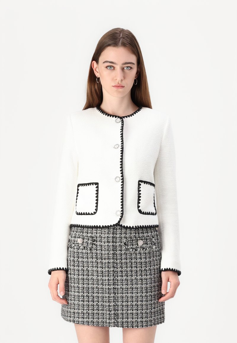 Veste blanche texturée avec des finitions noires, ornée de boutons ronds et de poches plaquées, associée à une jupe à carreaux noir et blanc.