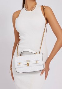 Biała torebka typu quilted z złotymi okuciami i górnym uchwytem. Wyposażona w łańcuszkowy pasek na ramię oraz zapięcie na klamrę. Widoczna nazwa marki "GUESS".