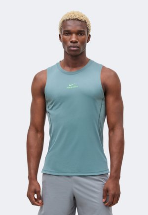 Hombre atlético con cabello rubio corto que lleva una camiseta sin mangas ajustada de Nike en color verde azulado y pantalones cortos gris claro, de pie frente a un fondo liso.