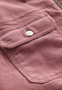 Gros plan sur une poche en tissu velours côtelé rose avec rabat et bouton argenté sur un vêtement, mettant en valeur la texture et les détails des coutures.