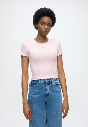 Kvinne med kort krøllete hår som har på seg en lys rosa tettsittende kortermet topp og høytlivs blå jeans mot en ensfarget bakgrunn.