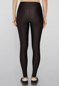 Leggings noirs avec une texture lisse et brillante et une taille haute, présentant un design ajusté qui se resserre jusqu'à la cheville, portés avec des chaussures décontractées.