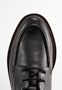 Chaussure en cuir noir avec une finition lisse, bout arrondi et accents cousus. Présente des lacets et une semelle brune slim.