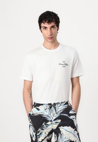 T-shirt bianco in cotone con maniche corte e scollo a girocollo, con un logo nero. Abbinato a pantaloni corti neri e blu con motivo a foglie.