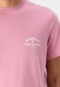 Světle růžové bavlněné tričko s kulatým výstřihem, krátkými rukávy a bílým potiskem loga, na kterém je napsáno "Barbour" a "A Life Outdoors."