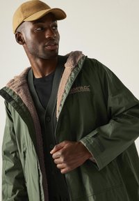 Man draagt een beige pet, groene jas met rits en fleece voering met het logo 'Regatta Great Outdoors', kijkt opzij.