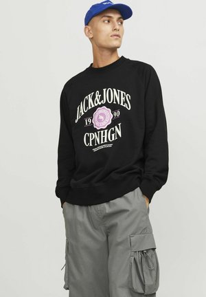Crni dukserica sa bijelim i ružičastim grafičkim tekstom "JACK&JONES", sivi cargo hlače s bočnim džepovima i plava kapa. Jednostavan dizajn, ležerna odjeća.