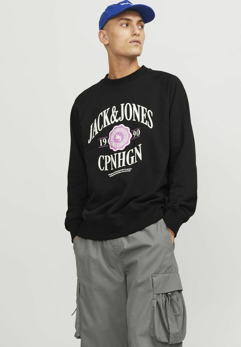 Sweatshirt noir avec le texte graphique blanc et rose "JACK&JONES", pantalon cargo gris avec poches latérales, et casquette bleue. Design simple, vêtements décontractés.