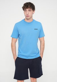 Ανοιχτό γαλάζιο βαμβακερό μπλουζάκι με στρογγυλή λαιμόκοψη και κοντά μανίκια. Διαθέτει μικρό μαύρο τυπωμένο λογότυπο στο στήθος. Φορεμένο με σκούρα σορτς.