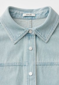 Col de veste en jean bleu clair avec trois boutons-pression argentés et une étiquette de marque "OPUS" à l'intérieur de l'encolure.