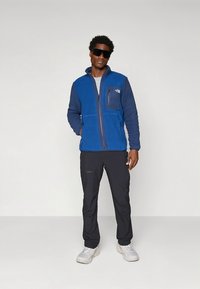 Μπλε φλις μπουφάν με φερμουάρ μπροστά, γκρίζο τσέπη, συνδυασμένο με σκούρο παντελόνι και λευκά παπούτσια, φορεμένο από άτομο με γυαλιά ηλίου σε ουδέτερο περιβάλλον.