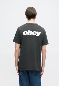 T-shirt cinza em algodão com mangas curtas, apresentando um grande gráfico branco com a inscrição "obey" nas costas. Ajuste casual, decote redondo básico e bainha não acabada.