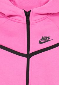 Różowa bluza z zamkiem Nike z czarnym zamkiem błyskawicznym, z czarnym logo oraz poziomymi czarnymi akcentami na ramionach. Miękka tekstura.